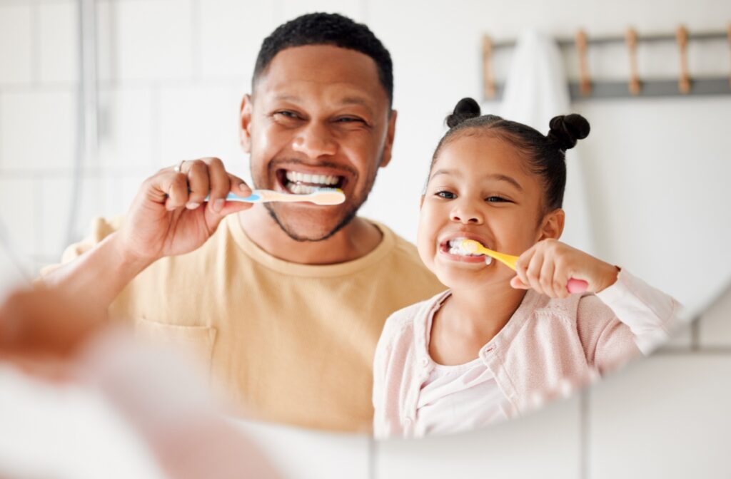 A parent and their child standing in front of a mirror while both brushing their teeth and smiling