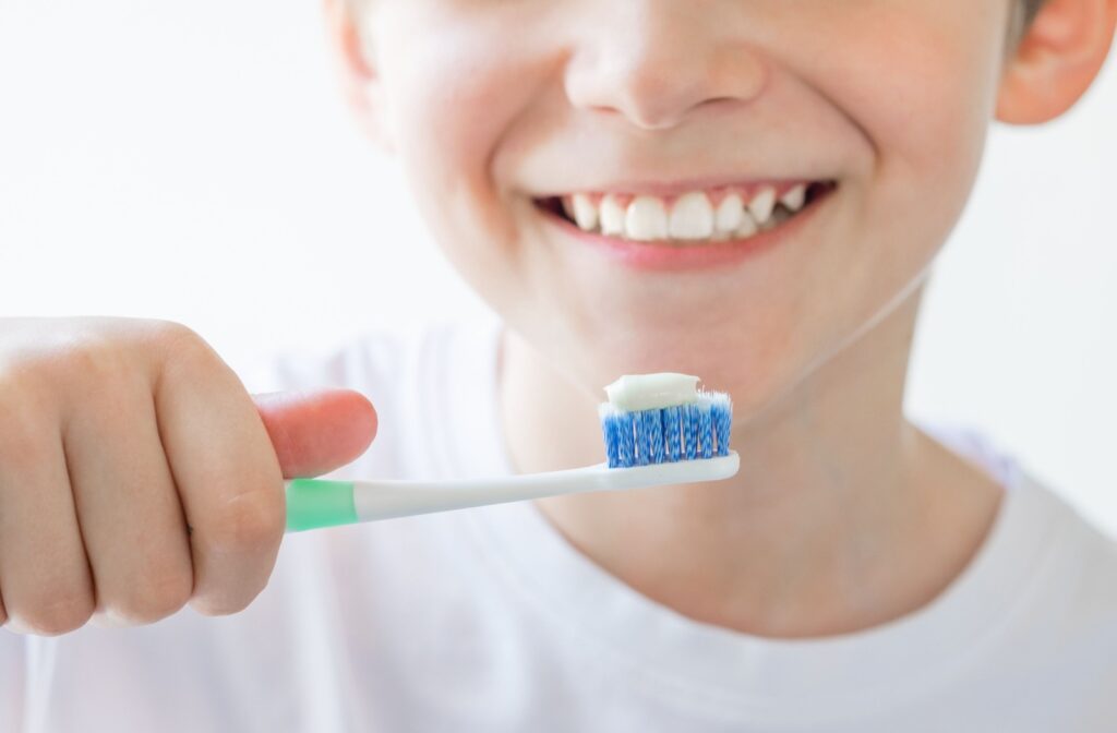 A child smiling at the camera before brushing his teeth, holding up a toothbrush with fluoride toothpaste.