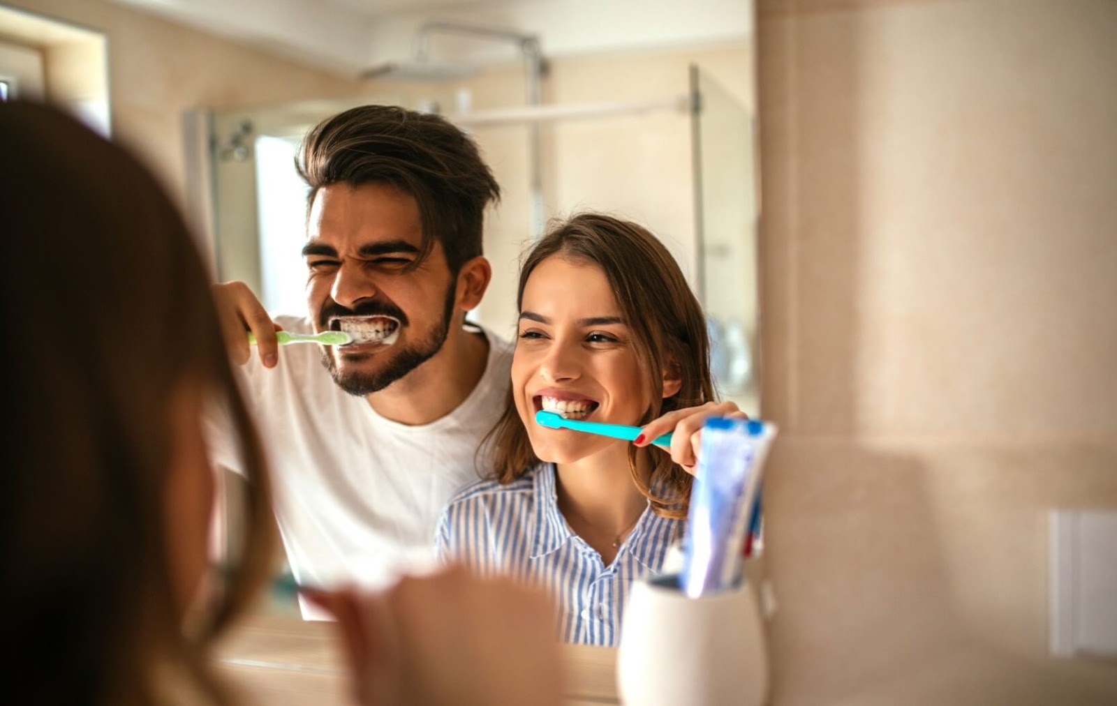 A happy young couple grin at each other in the mirror as they brush their teeth carefully side by side in a bathroom.