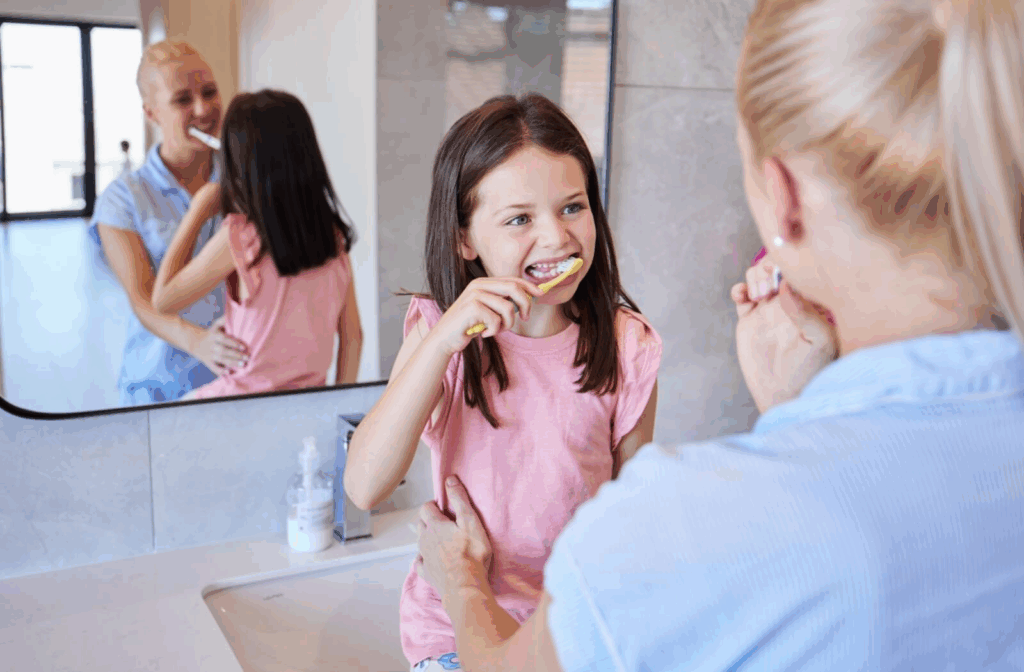 A child brushes their teeth at a bathroom sink while an adult brushes alongside them.