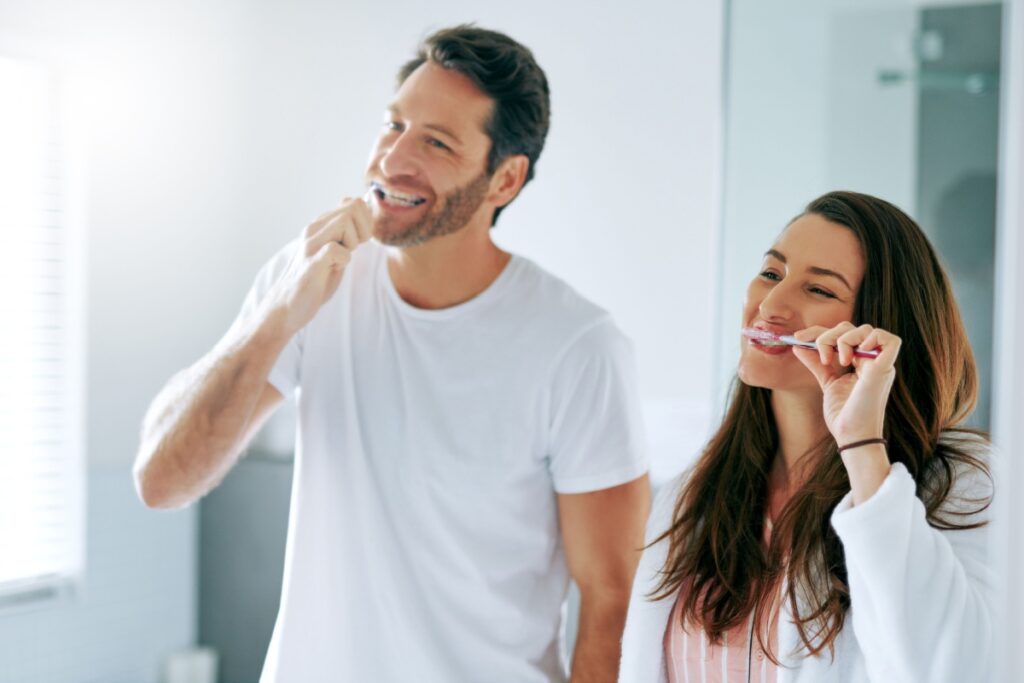 An adult couple brushing their teeth while looking in the bathroom mirror